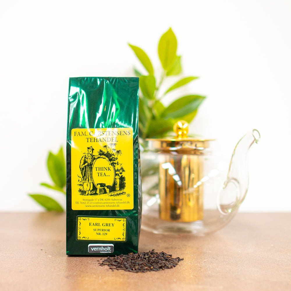 En mørkegrøn og gul pakke med teblanding fra Fam. Carstensens Tehandel. Varianten er med earl grey. I forgrunden ligger lidt løst te, og i baggrunden ses en tepotte af glas og gulddetaljer samt lidt grønne blade fra en plante.
