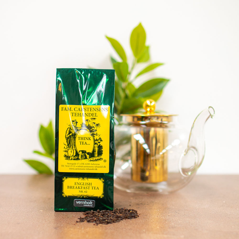 En mørkegrøn og gul pakke med teblanding fra Fam. Carstensens Tehandel. Varianten er med english breakfast tea. I forgrunden ligger lidt løst te, og i baggrunden ses en tepotte af glas og gulddetaljer samt lidt grønne blade fra en plante.