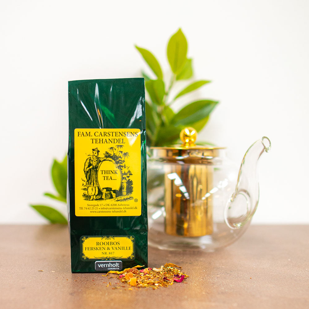 En mørkegrøn og gul pakke med teblanding fra Fam. Carstensens Tehandel. Varianten er med rooibos fersken og vanilje. I forgrunden ligger lidt løst te, og i baggrunden ses en tepotte af glas og gulddetaljer samt lidt grønne blade fra en plante.