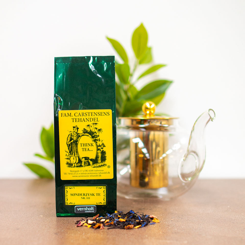 En mørkegrøn og gul pakke med teblanding fra Fam. Carstensens Tehandel. Varianten er med soenderjysk te. I forgrunden ligger lidt løst te, og i baggrunden ses en tepotte af glas og gulddetaljer samt lidt grønne blade fra en plante.
